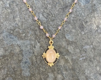 May include: A gold chain necklace with a pink oval gemstone pendant set in a gold filigree frame. The chain is made of small, round beads.
