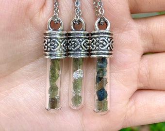 May include: Three silver necklaces with glass vials filled with small green, clear, and black stones. The vials have a silver cap with a decorative design.