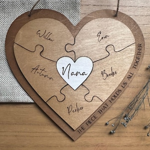 Puede incluir: Rompecabezas de madera en forma de coraz&oacute;n con la palabra "Nana" en un coraz&oacute;n blanco en el centro. Las piezas del rompecabezas est&aacute;n grabadas con los nombres: Willow, Autumn, Parker, Erin y Brooke. La frase "THE PIECE THAT HOLDS US ALL TOGETHER" est&aacute; escrita en el borde.