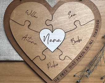 May include: Wooden heart-shaped puzzle with the word "Nana" in a white heart at the center. The puzzle pieces are engraved with names: Willow, Autumn, Parker, Erin, and Brooke. The phrase "THE PIECE THAT HOLDS US ALL TOGETHER" is written along the edge.