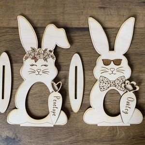 May include: Two wooden bunny-shaped Easter egg holders with slots for eggs. The bunny on the left has a floral crown and the text "Penelope" on the carrot. The bunny on the right has sunglasses, a bow tie, and the text "Oakley" on the carrot.