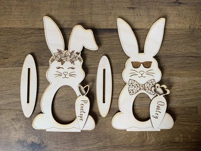 May include: Two wooden bunny-shaped Easter egg holders with slots for eggs. The bunny on the left has a floral crown and the text "Penelope" on the carrot. The bunny on the right has sunglasses, a bow tie, and the text "Oakley" on the carrot.