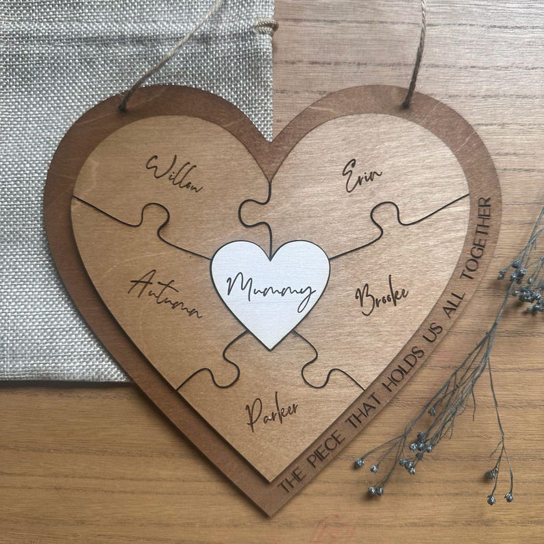 Puede incluir: Rompecabezas de madera en forma de coraz&oacute;n con la palabra "Mummy" en el centro. Las piezas del rompecabezas est&aacute;n grabadas con nombres y la frase "THE PIECE THAT HOLDS US ALL TOGETHER". Se adjunta una cuerda de yute para colgar.