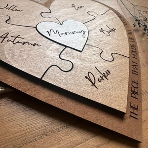 Puede incluir: Rompecabezas de madera en forma de coraz&oacute;n con la palabra "Mummy" en el centro. Las piezas del rompecabezas est&aacute;n grabadas con nombres. La frase "THE PIECE THAT HOLDS US ALL TOGETHER" est&aacute; impresa en el lateral. El rompecabezas es de madera marr&oacute;n claro.