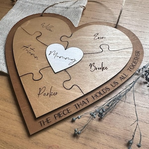 Puede incluir: Rompecabezas de madera en forma de coraz&oacute;n con cinco piezas, cada una con un nombre. Un peque&ntilde;o coraz&oacute;n blanco en el centro dice "Mummy". El borde exterior del rompecabezas dice "THE PIECE THAT HOLDS US ALL TOGETHER."
