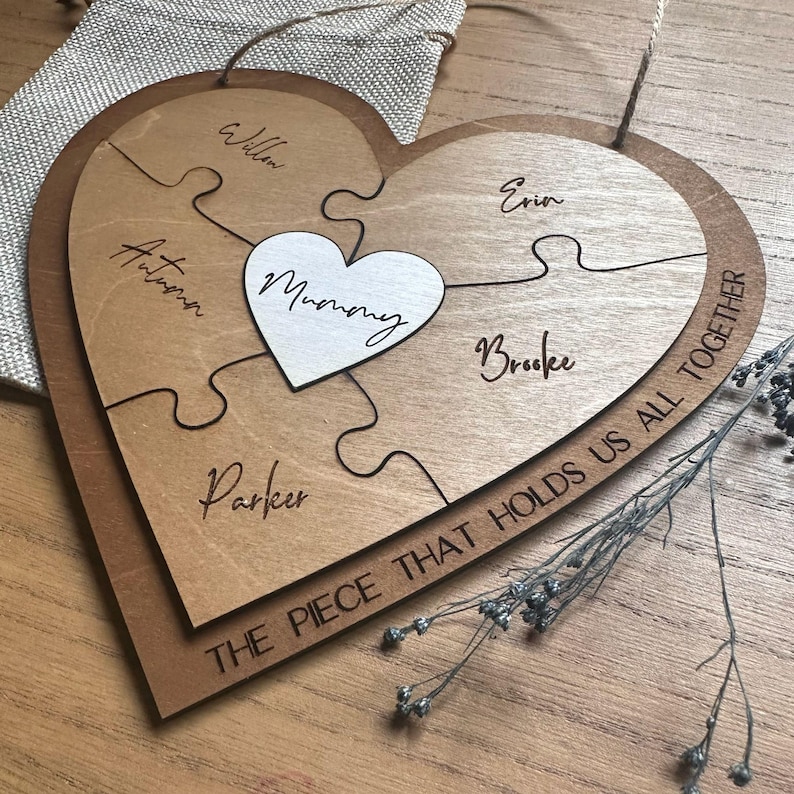 Puede incluir: Rompecabezas de madera en forma de coraz&oacute;n con cinco piezas, cada una con un nombre. Un peque&ntilde;o coraz&oacute;n blanco en el centro dice "Mummy". El borde exterior del rompecabezas dice "THE PIECE THAT HOLDS US ALL TOGETHER."