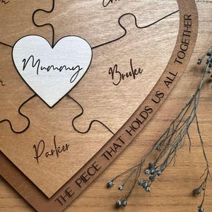 Puede incluir: Rompecabezas de madera en forma de coraz&oacute;n con la palabra "Mummy" en una pieza en forma de coraz&oacute;n blanca. Otras piezas tienen nombres como "Brooke" y "Parker". La frase "THE PIECE THAT HOLDS US ALL TOGETHER" est&aacute; grabada en una pieza curva.
