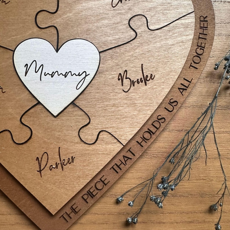Puede incluir: Rompecabezas de madera en forma de coraz&oacute;n con la palabra "Mummy" en una pieza en forma de coraz&oacute;n blanca. Otras piezas tienen nombres como "Brooke" y "Parker". La frase "THE PIECE THAT HOLDS US ALL TOGETHER" est&aacute; grabada en una pieza curva.