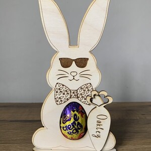 May include: Wooden Easter bunny cutout with sunglasses, a bow tie, and a carrot holding a chocolate egg. The carrot is personalized with the name "Oakley".