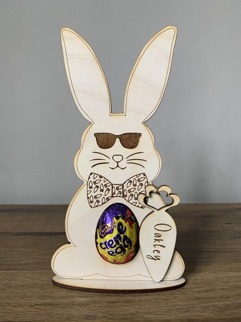 May include: Wooden Easter bunny cutout with sunglasses, a bow tie, and a carrot holding a chocolate egg. The carrot is personalized with the name "Oakley".