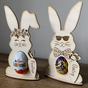 May include: Two wooden bunny rabbit Easter egg holders. The bunny on the left has a floral crown and is holding a Kinder Surprise egg. The bunny on the right is wearing sunglasses and a bow tie and is holding a Cadbury Creme Egg. Both bunnies have a carrot attached to the side with a name on it. The bunny on the left says "Penelope" and the bunny on the right says "Oakley".