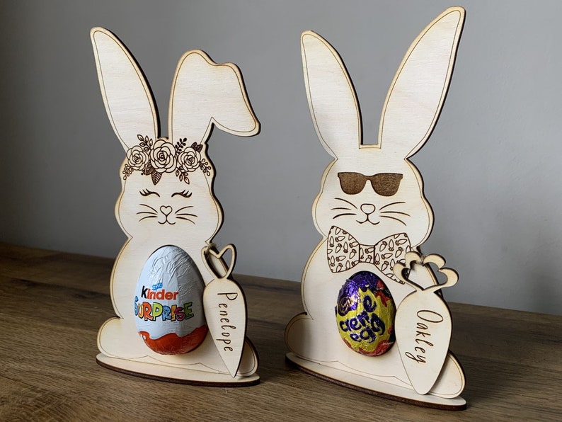 May include: Two wooden bunny rabbit Easter egg holders. The bunny on the left has a floral crown and is holding a Kinder Surprise egg. The bunny on the right is wearing sunglasses and a bow tie and is holding a Cadbury Creme Egg. Both bunnies have a carrot attached to the side with a name on it. The bunny on the left says "Penelope" and the bunny on the right says "Oakley".