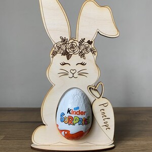 May include: Wooden bunny rabbit Easter egg holder with a floral crown, holding a Kinder Surprise egg. The bunny has a heart-shaped nose and the holder is personalized with the name "Penelope".