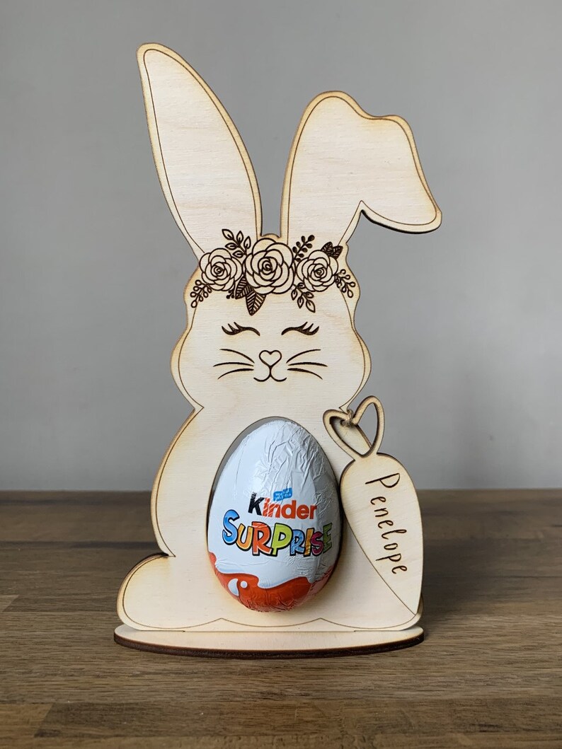 May include: Wooden bunny rabbit Easter egg holder with a floral crown, holding a Kinder Surprise egg. The bunny has a heart-shaped nose and the holder is personalized with the name "Penelope".