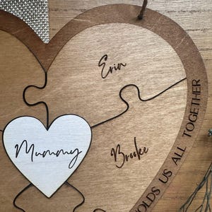 Puede incluir: Rompecabezas de madera en forma de coraz&oacute;n con cuatro piezas. La palabra "Mummy" est&aacute; en una pieza en forma de coraz&oacute;n blanca. Los nombres "Erin" y "Brooke" est&aacute;n grabados en las piezas de madera. La frase "HOLDS US ALL TOGETHER" est&aacute; grabada alrededor del borde.