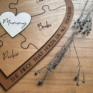 Puede incluir: Rompecabezas de madera con una pieza en forma de coraz&oacute;n que dice "Mummy". Otras piezas est&aacute;n grabadas con nombres. El rompecabezas est&aacute; inscrito con "THE PIECE THAT HOLDS US ALL TOGETHER". Flores secas sobre una superficie de madera.