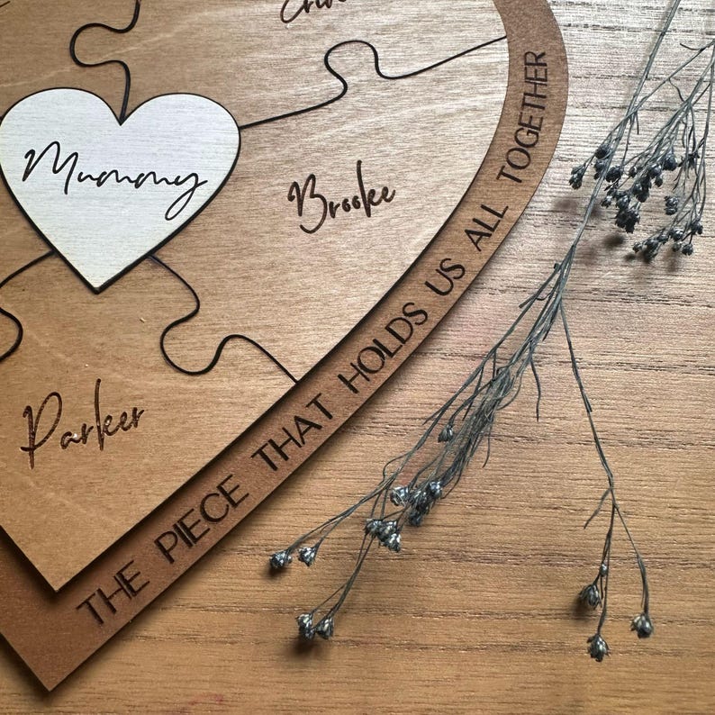Puede incluir: Rompecabezas de madera con una pieza en forma de coraz&oacute;n que dice "Mummy". Otras piezas est&aacute;n grabadas con nombres. El rompecabezas est&aacute; inscrito con "THE PIECE THAT HOLDS US ALL TOGETHER". Flores secas sobre una superficie de madera.