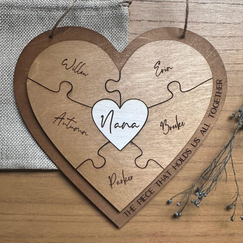 Puede incluir: Rompecabezas de madera en forma de coraz&oacute;n con la palabra "Nana" en un coraz&oacute;n blanco en el centro. Las piezas del rompecabezas est&aacute;n grabadas con los nombres: Willow, Autumn, Parker, Erin y Brooke. La frase "THE PIECE THAT HOLDS US ALL TOGETHER" est&aacute; escrita en el borde.