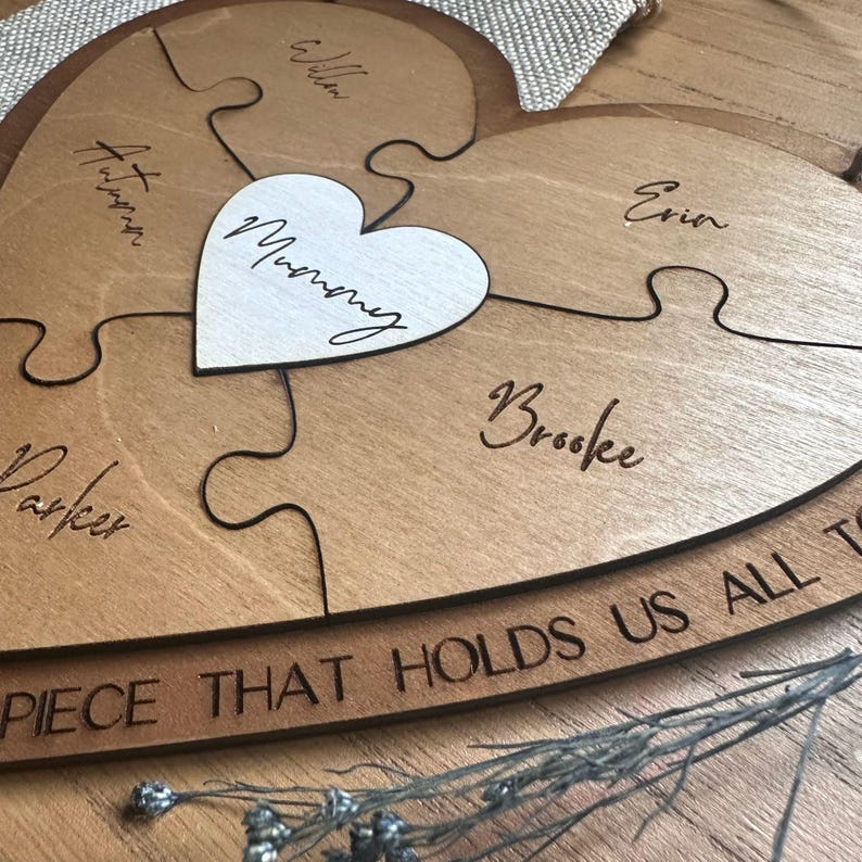 Puede incluir: Un rompecabezas de madera en forma de coraz&oacute;n con piezas individuales, cada una grabada con un nombre. Una pieza blanca en forma de coraz&oacute;n en el centro dice "Mummy". La frase "PIECE THAT HOLDS US ALL T" est&aacute; grabada en la parte inferior.