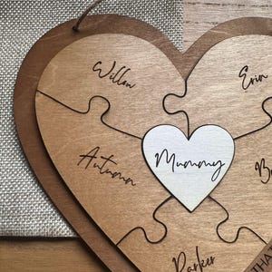 Puede incluir: Rompecabezas de madera en forma de coraz&oacute;n con acabado marr&oacute;n claro. El rompecabezas est&aacute; hecho de varias piezas, con la palabra "Mummy" en el centro. Las piezas individuales del rompecabezas est&aacute;n inscritas con nombres. Un art&iacute;culo decorativo.