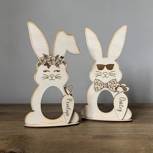 May include: Two wooden bunny egg holders, one with a floral crown and the name "Penelope" on a carrot, the other with sunglasses and a bow tie and the name "Oakley" on a carrot.