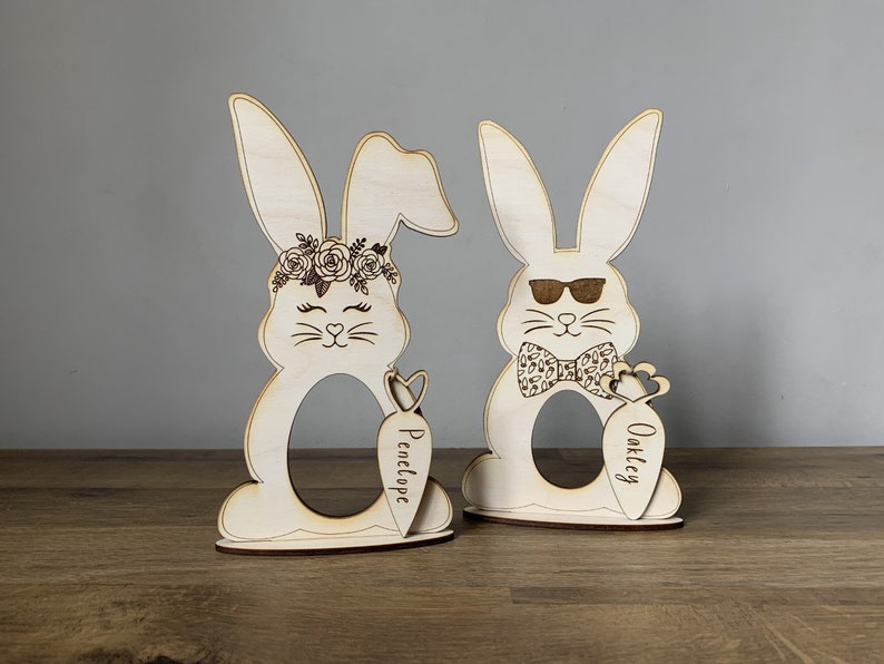 May include: Two wooden bunny egg holders, one with a floral crown and the name "Penelope" on a carrot, the other with sunglasses and a bow tie and the name "Oakley" on a carrot.