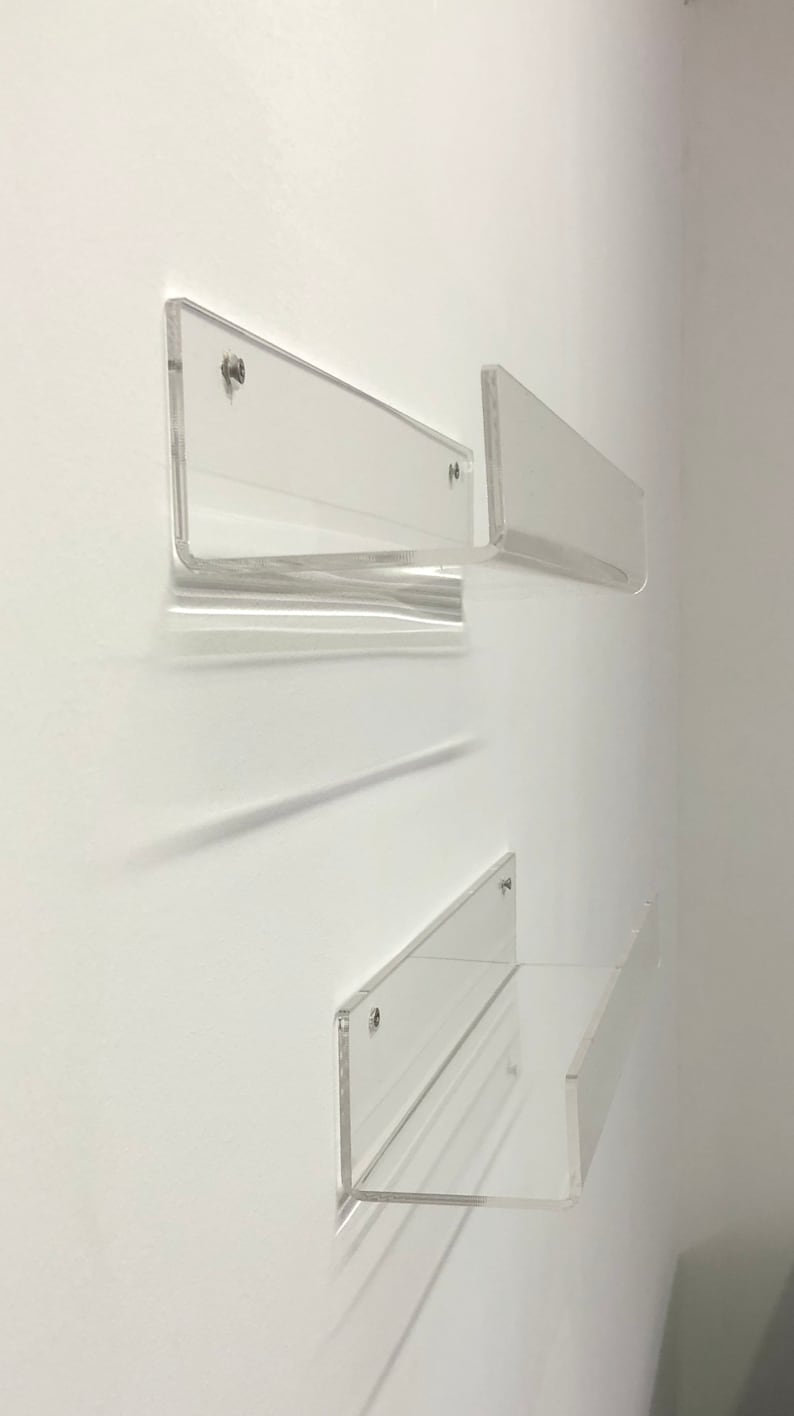 K&ouml;nnte beinhalten: Drei transparente Acrylregale an einer wei&szlig;en Wand montiert. Die Regale schweben und haben ein modernes, minimalistisches Design.