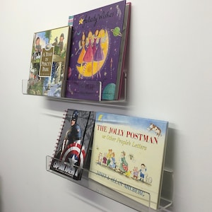 K&ouml;nnte beinhalten: Zwei transparente Acrylregale an einer wei&szlig;en Wand, die eine Auswahl an B&uuml;chern halten. Das obere Regal hat drei B&uuml;cher, eines mit einem violetten Einband und dem Titel "Felicity Wishes", ein anderes mit einem gr&uuml;nen Einband und dem Titel "A Year in Percy's Park" und ein drittes mit einem blauen Einband. Das untere Regal hat zwei B&uuml;cher, eines mit einem blauen Einband und dem Titel "The Jolly Postman or Other People's Letters" und ein anderes mit einem rot-wei&szlig;-blauen Einband und dem Titel "Captain America".