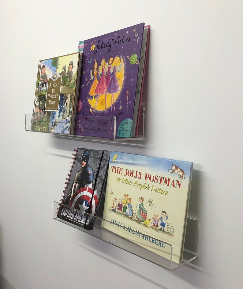 K&ouml;nnte beinhalten: Zwei transparente Acrylregale an einer wei&szlig;en Wand, die eine Auswahl an B&uuml;chern halten. Das obere Regal hat drei B&uuml;cher, eines mit einem violetten Einband und dem Titel "Felicity Wishes", ein anderes mit einem gr&uuml;nen Einband und dem Titel "A Year in Percy's Park" und ein drittes mit einem blauen Einband. Das untere Regal hat zwei B&uuml;cher, eines mit einem blauen Einband und dem Titel "The Jolly Postman or Other People's Letters" und ein anderes mit einem rot-wei&szlig;-blauen Einband und dem Titel "Captain America".