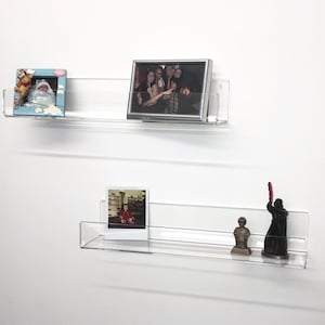K&ouml;nnte beinhalten: Zwei transparente Acrylregale, die an einer wei&szlig;en Wand montiert sind, jedes mit gerahmten Fotos und kleinen Figuren. Das obere Regal hat ein gerahmtes Foto einer Gruppe von Personen und ein kleineres gerahmtes Foto eines Babys. Das untere Regal hat ein gerahmtes Foto eines Kindes, eine kleine Statue einer Person und eine schwarze Figur einer Person, die ein rotes Lichtschwert h&auml;lt.