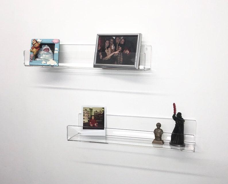 K&ouml;nnte beinhalten: Zwei transparente Acrylregale, die an einer wei&szlig;en Wand montiert sind, jedes mit gerahmten Fotos und kleinen Figuren. Das obere Regal hat ein gerahmtes Foto einer Gruppe von Personen und ein kleineres gerahmtes Foto eines Babys. Das untere Regal hat ein gerahmtes Foto eines Kindes, eine kleine Statue einer Person und eine schwarze Figur einer Person, die ein rotes Lichtschwert h&auml;lt.
