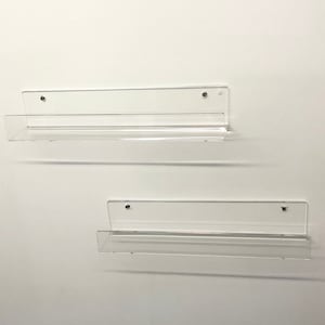 K&ouml;nnte beinhalten: Zwei transparente Acrylregale, die an einer wei&szlig;en Wand montiert sind. Die Regale sind f&uuml;r die Aufbewahrung von kleinen Gegenst&auml;nden wie B&uuml;chern, Schmuck oder anderen Dekorationsartikeln konzipiert.