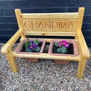 May include: A wooden bench with the word "GRANDMA" carved into the back. The bench has two planter boxes on the front, each with a purple flower.