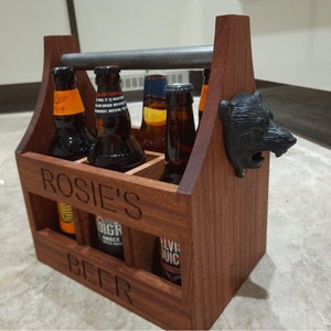 May include: A wooden beer caddy holding six beer bottles. The caddy is made of dark wood and has a metal handle. The words "Rosie's Beer" are carved into the front. A black bear head bottle opener is attached to the side.