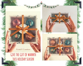 May include: A white marble gift box with a gold ribbon and four round, colorful bath bombs. The box is held by hands and the text "Give the gift of warmth this holiday season" is printed below the box.  Kristal K Creations is printed above the box.