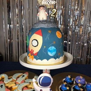 May include: A blue and white astronaut cake with a rocket, planets, and stars. The cake is decorated with a small astronaut figurine and a gold number 2. The cake is on a white cake stand. There are also cupcakes and cookies decorated with a space theme.