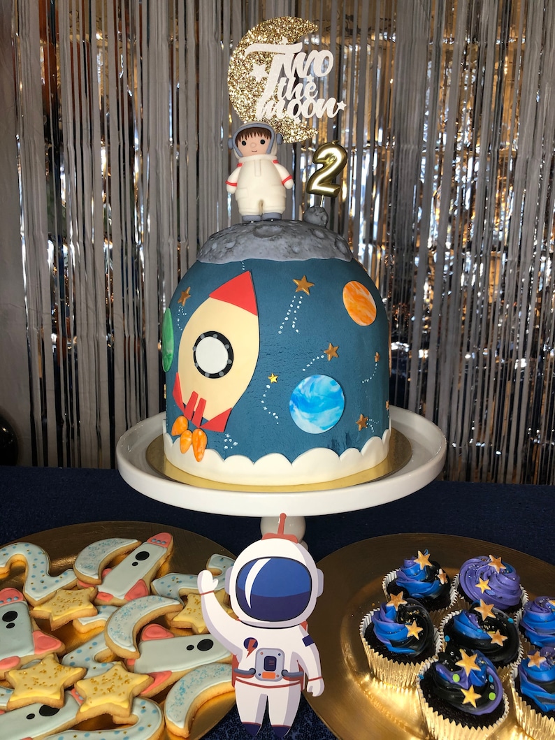 May include: A blue and white astronaut cake with a rocket, planets, and stars. The cake is decorated with a small astronaut figurine and a gold number 2. The cake is on a white cake stand. There are also cupcakes and cookies decorated with a space theme.