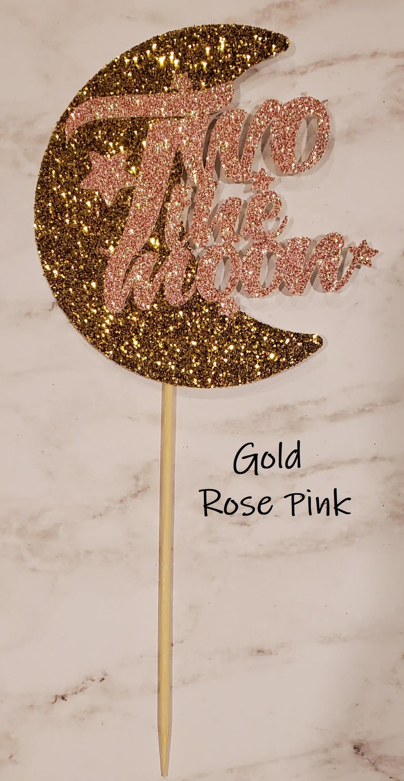May include: Gold glitter crescent moon cake topper with the words "Two the Moon" in rose pink glitter.