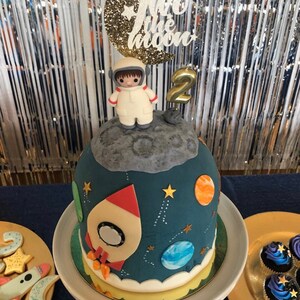 May include: A blue and white space-themed cake with a rocket, planets, and stars. The cake is decorated with a figurine of an astronaut standing on the moon. The cake topper says "Two the Moon" and has a gold glitter finish.