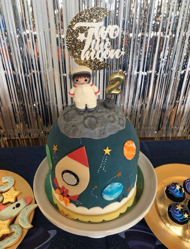 May include: A blue and white space-themed cake with a rocket, planets, and stars. The cake is decorated with a figurine of an astronaut standing on the moon. The cake topper says "Two the Moon" and has a gold glitter finish.