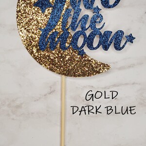 May include: Gold glitter crescent moon cake topper with the words "Two the Moon" in dark blue glitter.