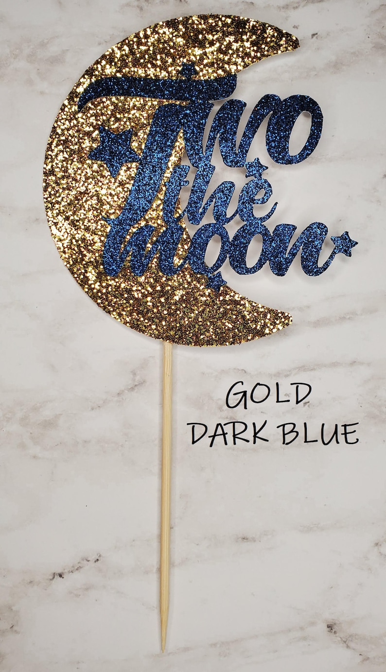 May include: Gold glitter crescent moon cake topper with the words "Two the Moon" in dark blue glitter.