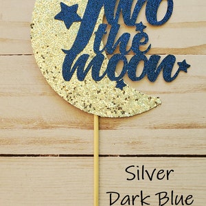 May include: A gold glitter crescent moon with a dark blue glitter "Two the Moon" cake topper.