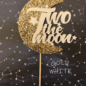 May include: Gold glitter moon cake topper with the words "Two the Moon" in white. The topper is on a black background with white stars.
