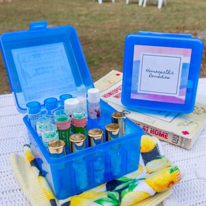 May include: A blue plastic container with a label that says "Homeopathic Remedies" and is filled with small glass bottles. The container is sitting on a white and yellow striped towel.