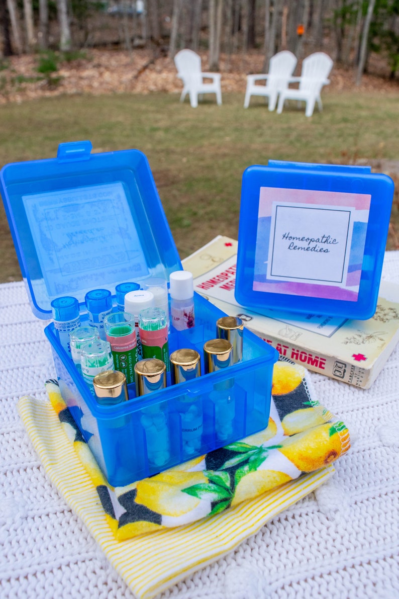 May include: A blue plastic container with a label that says "Homeopathic Remedies" and is filled with small glass bottles. The container is sitting on a white and yellow striped towel.