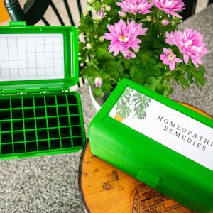 May include: Two green plastic storage boxes, one open and one closed, labelled "HOMEOPATHIC REMEDIES". The open box has a grid of small compartments. The boxes are on a wooden surface, with pink flowers in the background.