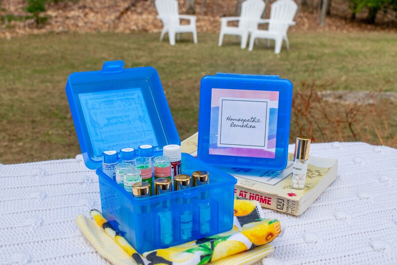 May include: A blue plastic box with a label that says "Homeopathic Remedies" and is filled with small glass bottles. The box is sitting on a white crocheted blanket with a yellow and white patterned towel.