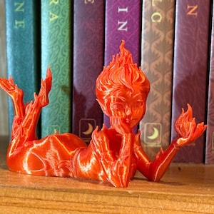 May include: A 3D-printed orange figurine of a reclining humanoid figure with flames for hair and limbs. The figure is resting on a wooden shelf in front of a row of colorful books. The figurine is a unique decorative item.