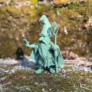 Puede incluir: Una figurita verde de un mago con una larga barba y un sombrero puntiagudo, sosteniendo un bastón con una parte superior decorativa. La figurita está de pie sobre un lecho de musgo.